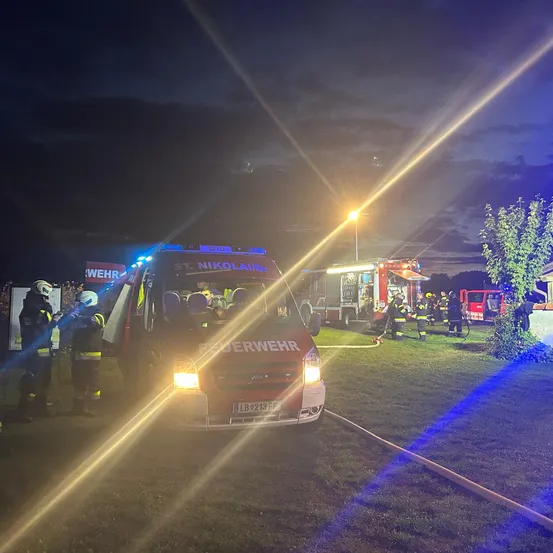 Feuerwehrleute in Schutzkleidung arbeiten bei Nacht. Ein roter Feuerwehrwagen ist mit eingeschalteten Lichtern geparkt. Ein Feuerwehrauto ist ebenfalls in der Nähe geparkt.