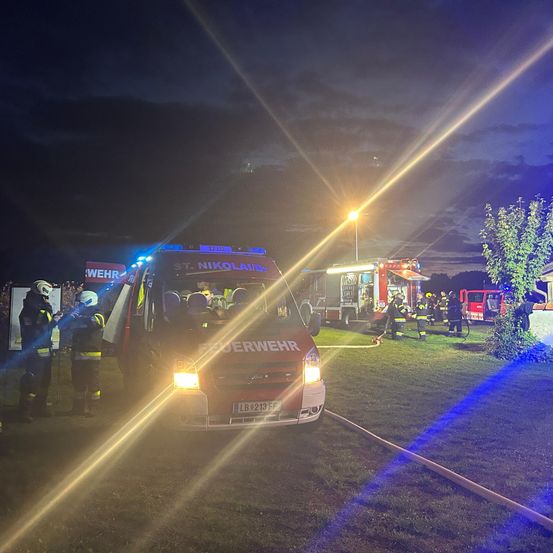 Feuerwehrleute in Schutzkleidung arbeiten bei Nacht. Ein roter Feuerwehrwagen ist mit eingeschalteten Lichtern geparkt. Ein Feuerwehrauto ist ebenfalls in der Nähe geparkt.