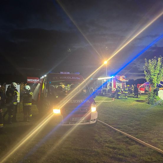 In der Nacht steht ein Feuerwehrwagen mit der Aufschrift St. Nikolai/Dr. auf einem Rasen. Feuerwehrleute stehen um ihn herum, und im Hintergrund befinden sich Zelte und ein Feuerwehrwagen.