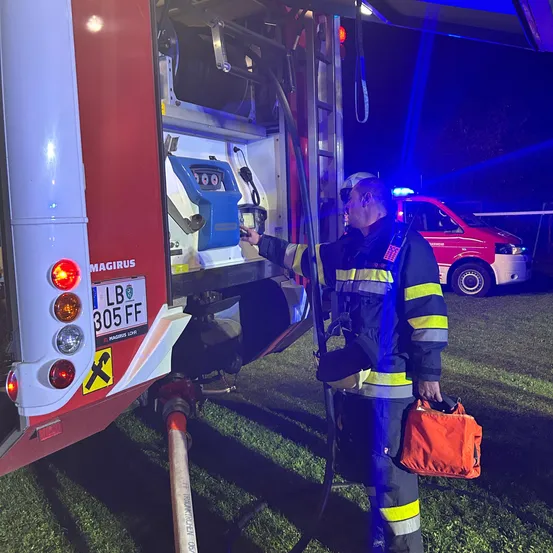 Ein Feuerwehrmann mit einer roten Tasche steht neben einem Feuerwehrwagen, dessen Tür offen ist. Der Wagen hat ein Schild mit LB 305 FF. Ein roter Van ist dahinter geparkt.