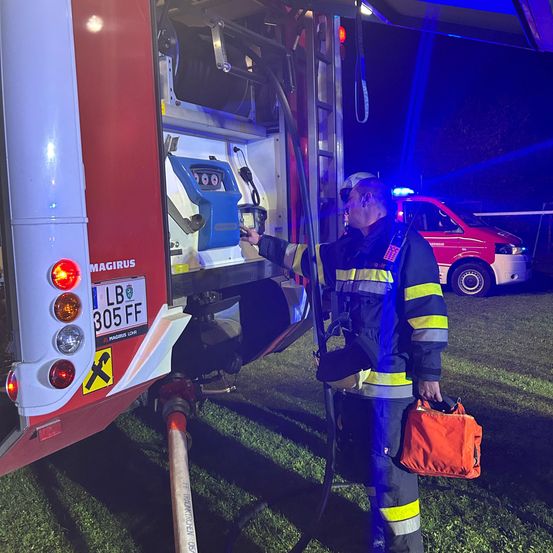 Ein Feuerwehrmann mit einer roten Tasche steht neben einem Feuerwehrwagen, dessen Tür offen ist. Der Wagen hat ein Schild mit LB 305 FF. Ein roter Van ist dahinter geparkt.