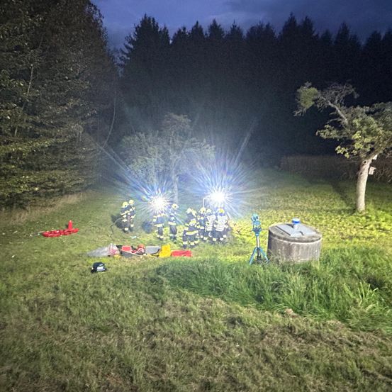 Eine nächtliche Szene mit einer Gruppe von Feuerwehrleuten, die in einem Grasbereich mit Taschenlampen umgeben von Bäumen stehen.