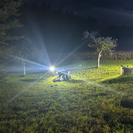 In der Nacht wird ein Feld von einem hellen Licht beleuchtet. Eine Person liegt mit einem Rucksack in der Nähe auf dem Gras. Auf beiden Seiten des Feldes stehen zwei Bäume.