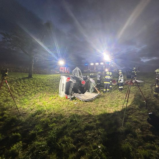 Ein Unfallort in der Nacht mit anwesenden Rettungsdiensten, das Fahrzeug liegt auf dem Kopf auf einem Grasfeld, während Rettungslichter den Bereich beleuchten.