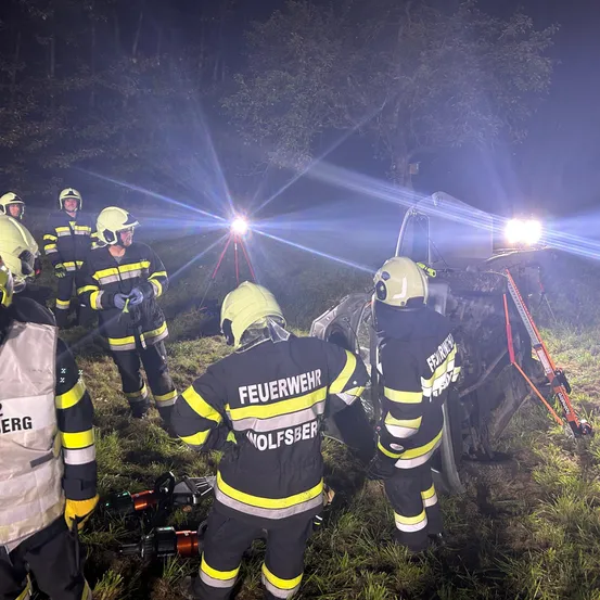 Feuerwehrleute arbeiten nachts daran, ein Auto aus einer Schlucht zu befreien. Sie sind mit Helmen, Handschuhen und Schutzanzügen ausgestattet. Sie benutzen Werkzeuge und eine Winde. Das Auto ist von Gras und Bäumen umgeben.