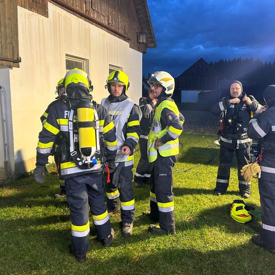 Eine Gruppe von Feuerwehrleuten ist nachts vor einem Gebäude versammelt. Sie tragen gelbe und schwarze Uniformen und Helme. Einer von ihnen hat eine gelbe Sauerstoffflasche auf dem Rücken. Ein gelber Helm liegt in der Nähe auf dem Boden. Der Bereich wird vom Licht des Gebäudes beleuchtet.
