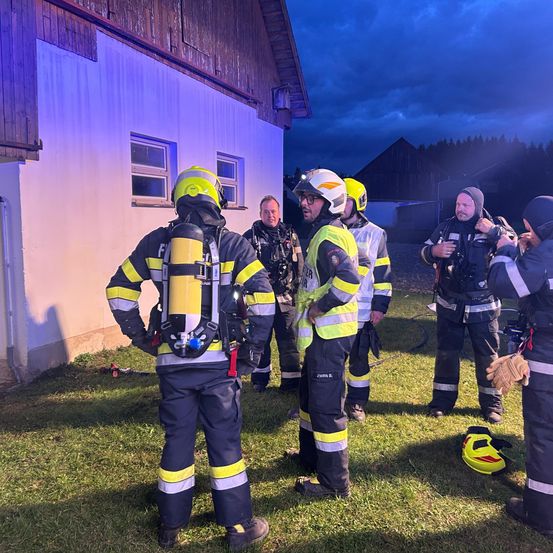 Feuerwehrleute versammeln sich vor einem Gebäude und besprechen Strategien. Eine bewölkte Nacht mit schwacher Beleuchtung. Helme und Sicherheitsausrüstung sind angelegt.