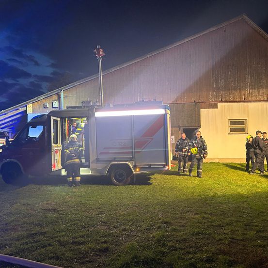 Feuerwehrleute stehen vor einem brennenden Feuerwehrwagen, der von seinem eigenen Licht beleuchtet wird, vor einem Gebäude unter einem bewölkten Nachthimmel.