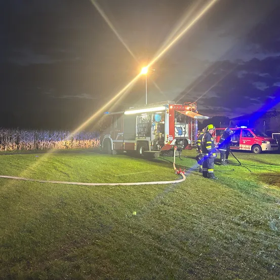 Feuerwehrleute sind nachts auf einem Rasenfeld und benutzen einen Schlauch von einem Feuerwehrwagen. Blaue Lichter beleuchten die Szene.