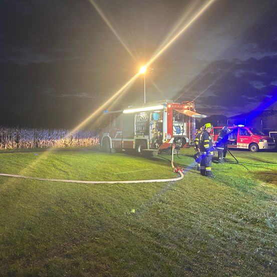 Feuerwehrleute sind nachts auf einem Rasenfeld und benutzen einen Schlauch von einem Feuerwehrwagen. Blaue Lichter beleuchten die Szene.