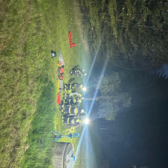 Eine Nachtszene zeigt Feuerwehrleute in voller Ausrüstung mit Helmen und Taschenlampen, die eine rote Trage umgeben. Der Bereich ist durch ihre Lichter beleuchtet, mit hohen Bäumen im Hintergrund.