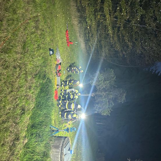 Eine Nachtszene zeigt Feuerwehrleute in voller Ausrüstung mit Helmen und Taschenlampen, die eine rote Trage umgeben. Der Bereich ist durch ihre Lichter beleuchtet, mit hohen Bäumen im Hintergrund.