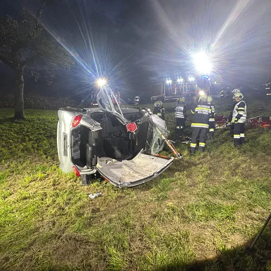 Ein Auto ist auf einem Grasfeld bei Nacht umgekippt. Rettungskräfte kümmern sich darum, und ihre Lichter beleuchten die Szene.