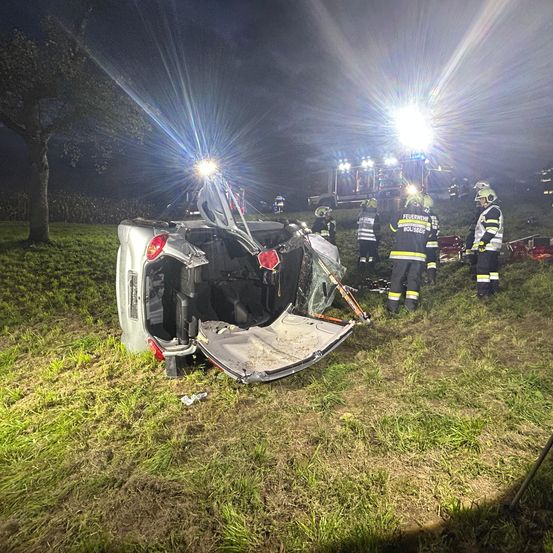 Ein Auto ist auf einem Grasfeld bei Nacht umgekippt. Rettungskräfte kümmern sich darum, und ihre Lichter beleuchten die Szene.