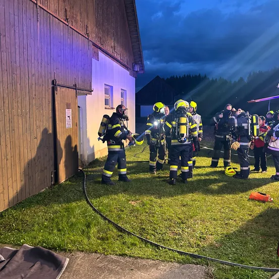 Feuerwehrleute stehen bei Nacht vor einem Gebäude, tragen Helme und Schutzausrüstung. Einige halten Taschenlampen.