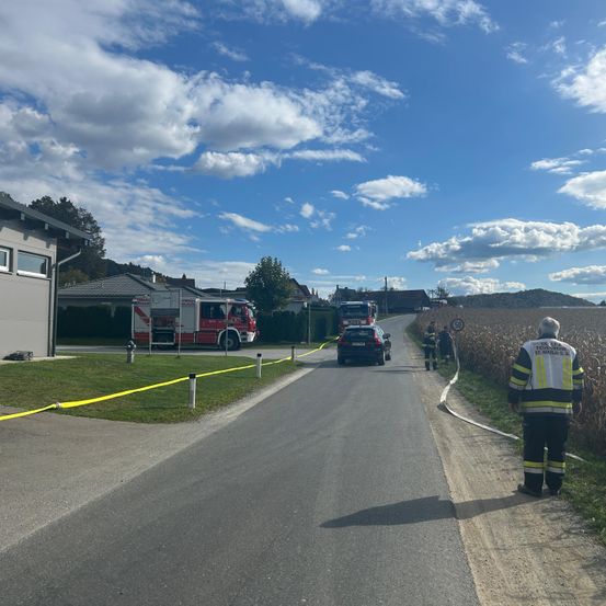 Ein Feuerwehrauto ist an einer Straße mit einem gelben Seil geparkt, und eine Person in einer Feuerwehruniform steht auf der Seite. Zwei Personen stehen mit einer Feuerwehrschlauch auf der Straße, und ein weiteres Auto ist in der Nähe geparkt.