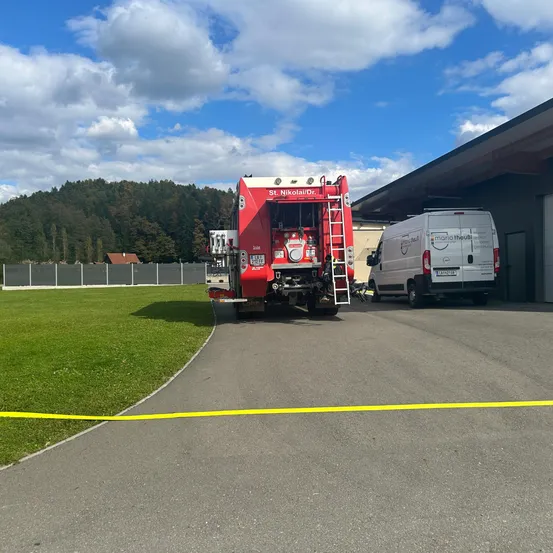 Ein roter Feuerwehrwagen mit Leiter steht vor einem Gebäude, neben dem ein weißer Lieferwagen geparkt ist. Ein gelbes Warnband liegt auf dem Boden.