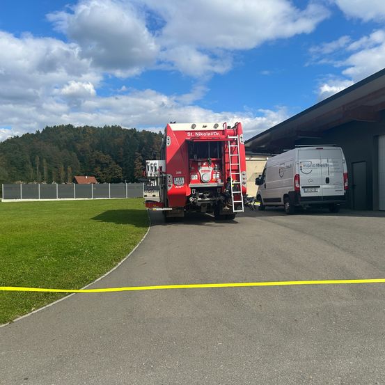 Ein roter Feuerwehrwagen mit Leiter steht vor einem Gebäude, neben dem ein weißer Lieferwagen geparkt ist. Ein gelbes Warnband liegt auf dem Boden.