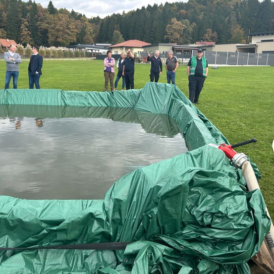 Eine Gruppe von Menschen steht um einen großen mit grünem Planen bedeckten Wasserpool herum, der sich auf einem Rasenfeld mit Bäumen im Hintergrund befindet.