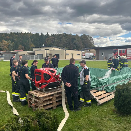 Eine Gruppe von Feuerwehrleuten versammelt sich auf einem Rasenfeld. Sie üben mit einem roten Feuerwehrauto. Das Gebiet ist von Büschen und Bäumen umgeben.