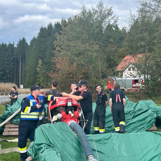 Eine Gruppe von Feuerwehrleuten in Uniform arbeitet auf einem Grasfeld mit grünen Planen an einem Fahrzeug. Bäume und ein Gebäude sind im Hintergrund zu sehen.