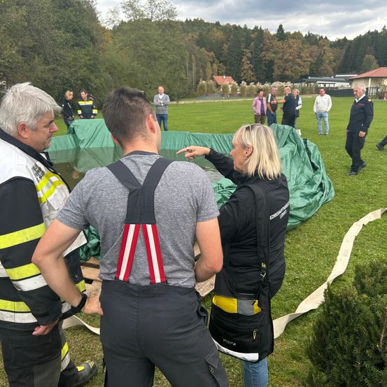 Eine Gruppe von Menschen auf einem Rasenfeld. Ein Mann in einer Hose mit Aufhängung und eine Frau in einer schwarzen Jacke betrachten einen abgedeckten Pool. Andere Personen unterhalten sich im Hintergrund.