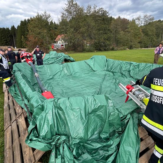 Eine Gruppe Feuerwehrleute in Uniform arbeitet an einer großen grünen Plane, neben einem roten Fahrzeug, in einem Grasfeld mit Bäumen und einem Haus im Hintergrund.