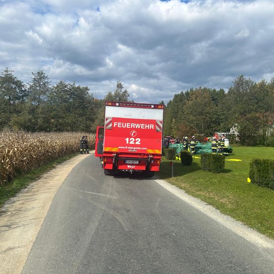 Ein Feuerwehrauto steht am Straßenrand, daneben stehen Feuerwehrleute. Auf der Wiese sind grüne Zelte aufgebaut, links ist Mais gepflanzt.