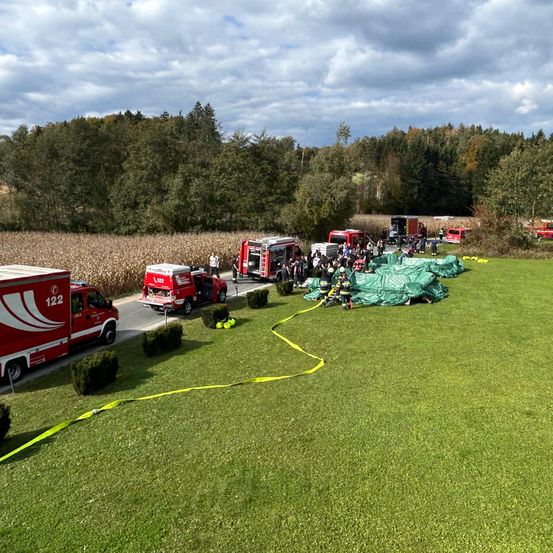 Rettungsfahrzeuge umgeben eine grüne Plane auf einem Rasenfeld mit einem Maisfeld und Bäumen im Hintergrund.
