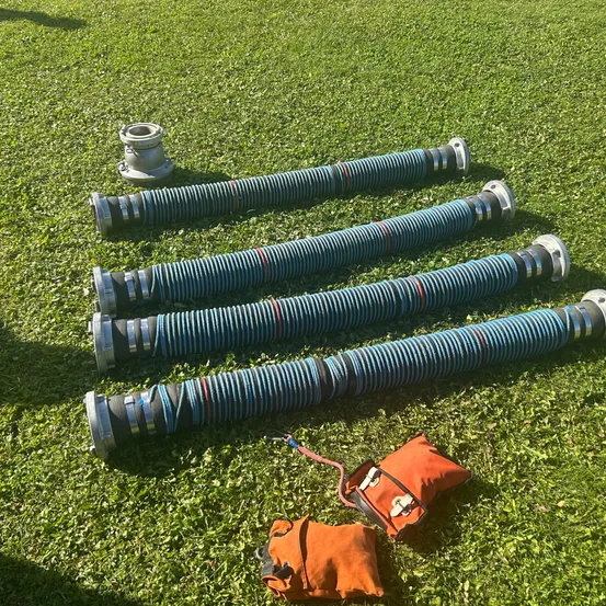 Fünf lange blaue Schläuche liegen auf grünem Gras mit zwei orangefarbenen Beuteln in der Nähe. Die Schläuche sind aufgerollt und haben silberne Enden. Die Beutel sind durch ein rotes Kabel verbunden.