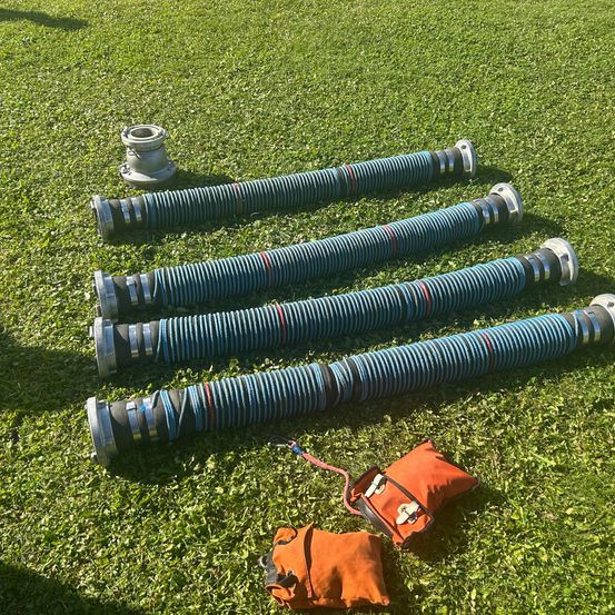 Fünf lange blaue Schläuche liegen auf grünem Gras mit zwei orangefarbenen Beuteln in der Nähe. Die Schläuche sind aufgerollt und haben silberne Enden. Die Beutel sind durch ein rotes Kabel verbunden.