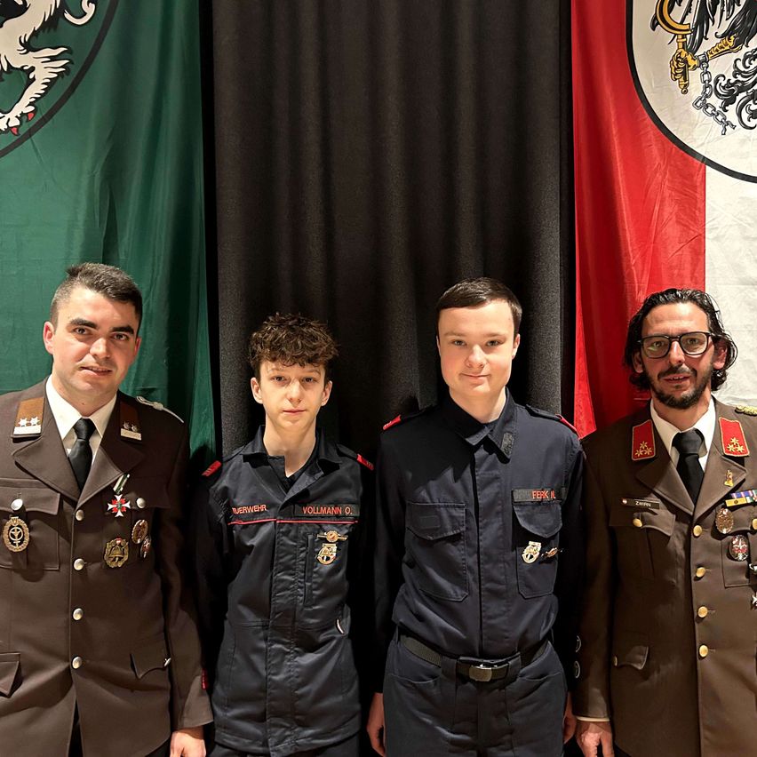 Vier Männer in Uniformen stehen vor verschiedenen Flaggen. Der Mann in der Mitte hat 'VOLLMANN O.' auf seiner Jacke.