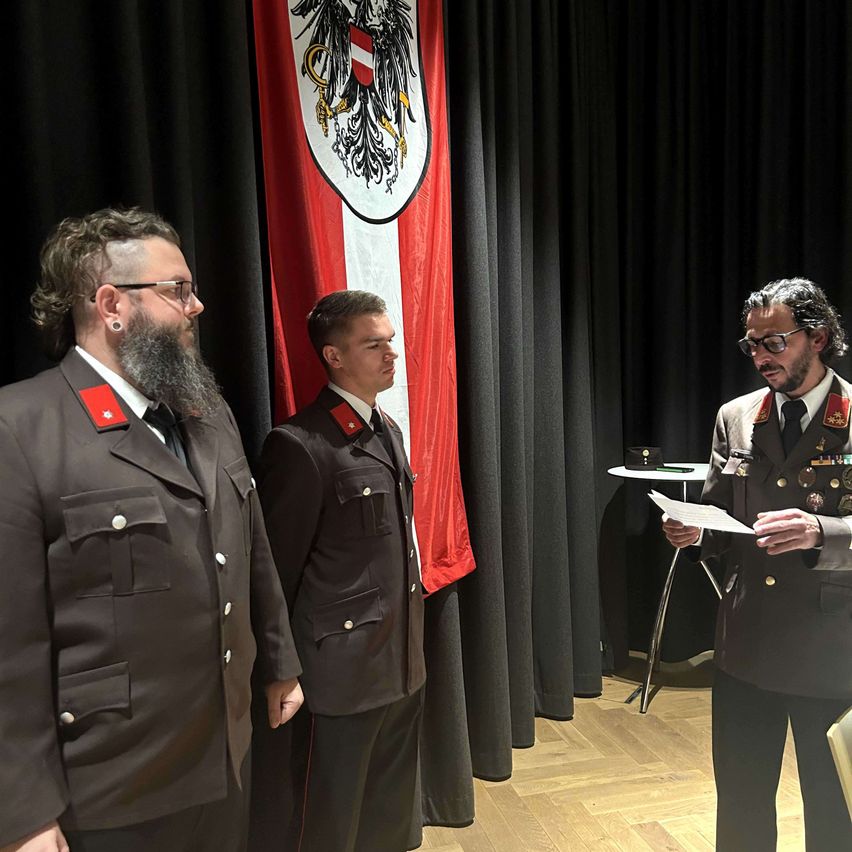 Drei Männer in Militäruniformen stehen neben einer Flagge mit einem Wappen. Einer liest ein Dokument, ein anderer schaut ihn an, und der dritte schaut weg. Hinter ihnen steht ein Tisch mit Gegenständen.