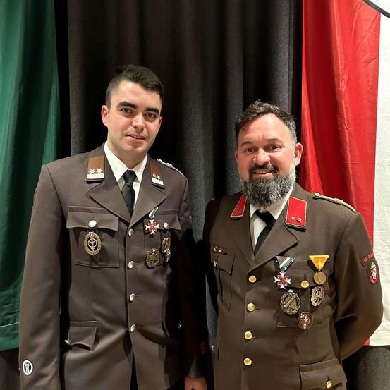 Zwei Männer in Militäruniformen stehen nebeneinander vor Flaggen. Sie haben beide Medaillen an ihren Uniformen.