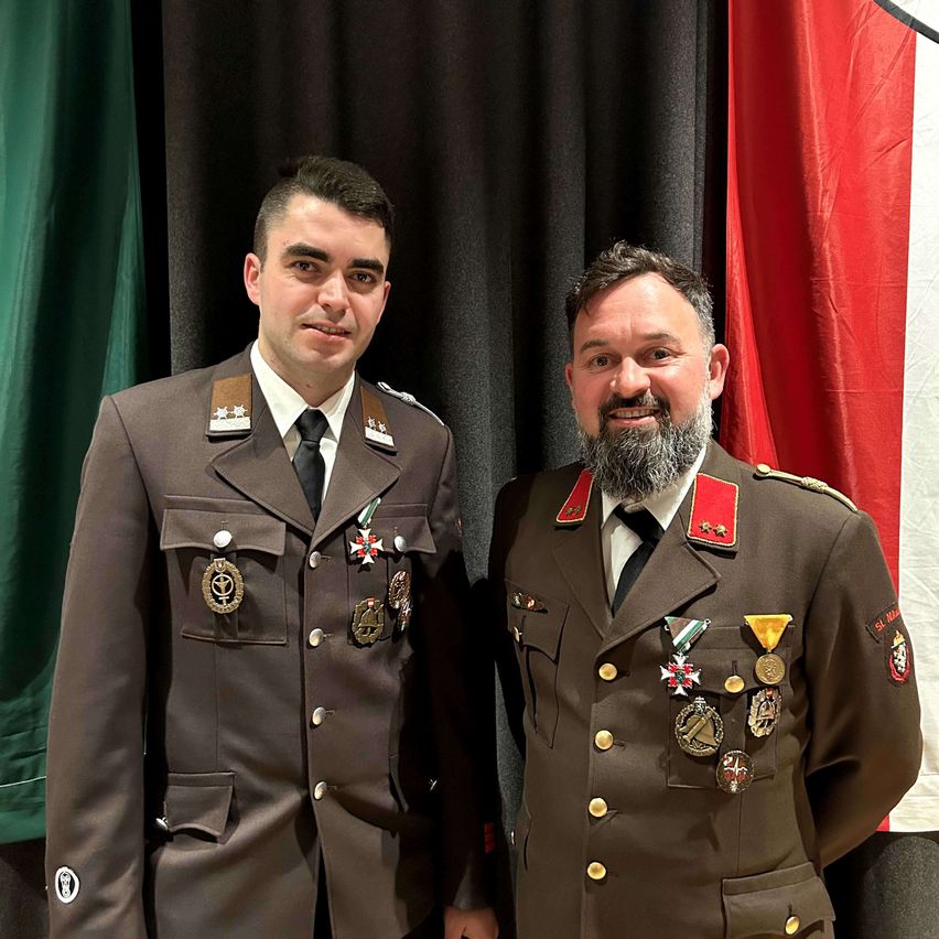 Zwei Männer in Militäruniformen stehen nebeneinander vor Flaggen. Sie haben beide Medaillen an ihren Uniformen.