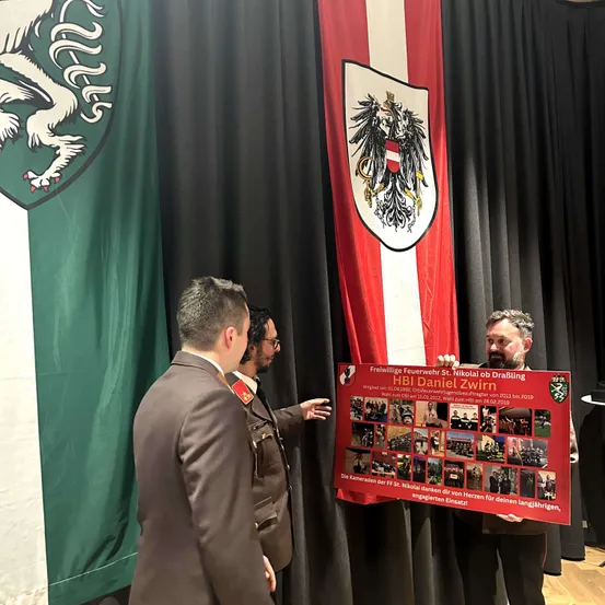 Drei Männer in Uniformen stehen vor einem roten Banner. Sie sind bei einer Veranstaltung, mit einer grünen Flagge und schwarzen Vorhängen dahinter. Das Banner zeigt eine Fotocollage und Text über HBI Daniel Zwirn.