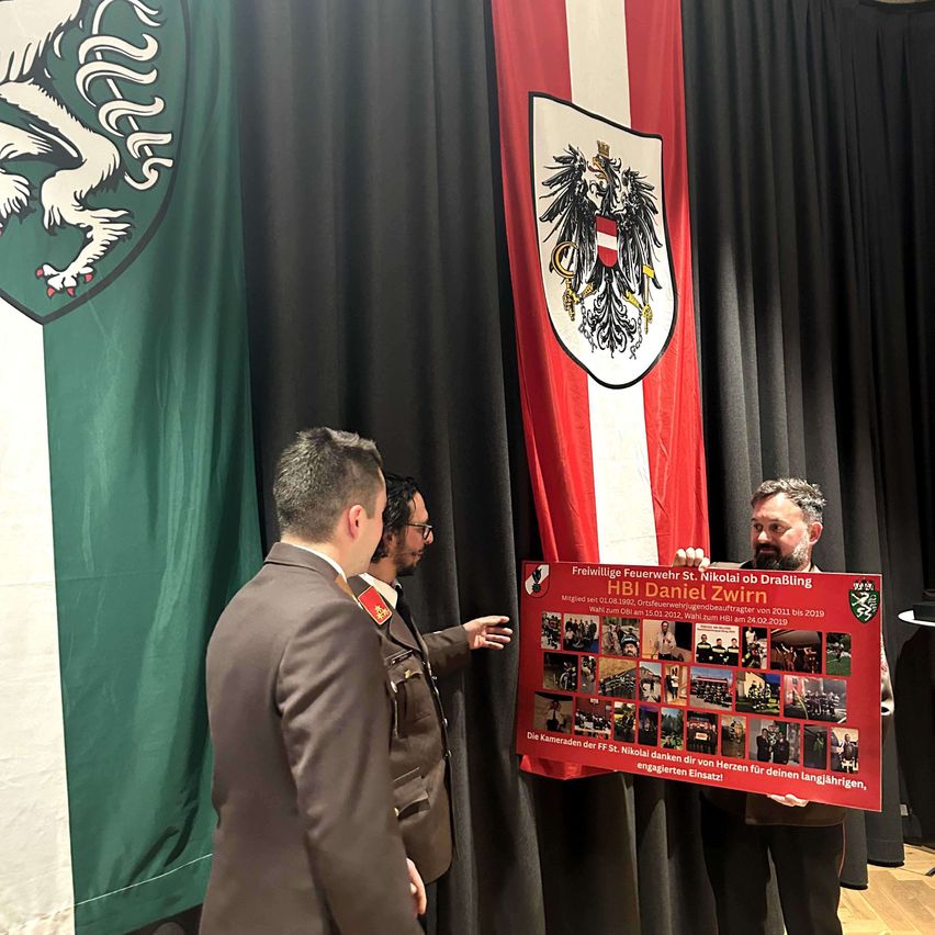 Drei Männer in Uniformen stehen vor einem roten Banner. Sie sind bei einer Veranstaltung, mit einer grünen Flagge und schwarzen Vorhängen dahinter. Das Banner zeigt eine Fotocollage und Text über HBI Daniel Zwirn.