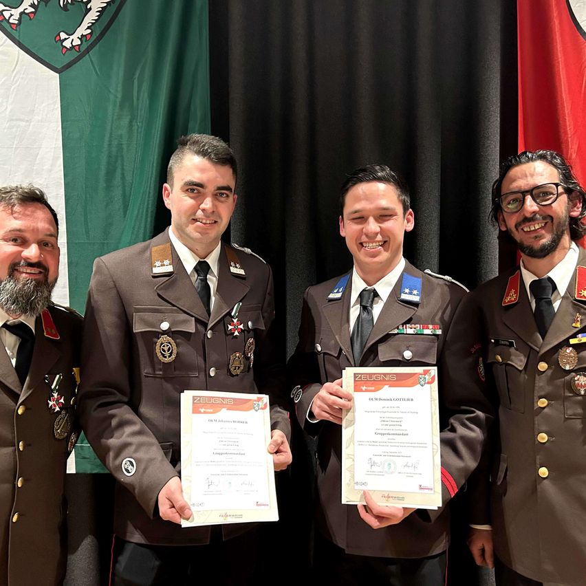 Vier Männer in Militäruniformen stehen vor einer Fahne und lächeln für ein Foto. Zwei Männer halten Urkunden mit Text und Logos.