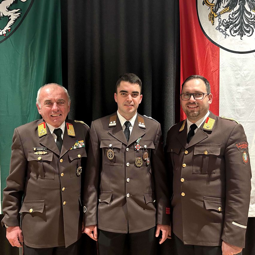 Drei Männer in Militäruniformen stehen vor einer Kulisse mit Fahnen. Der Mann auf der linken Seite hat Medaillen auf seiner linken Brust.