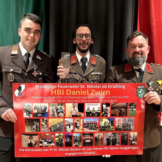 Drei Männer in Militäruniformen stehen zusammen und halten ein rotes Banner mit der Aufschrift 'Freiwillige Feuerwehr St. Nikolai ob Drablig HBI Daniel Zwirn'. Das Banner zeigt Fotos von Menschen und Aktivitäten. Der Mann in der Mitte hält eine Trophäe.