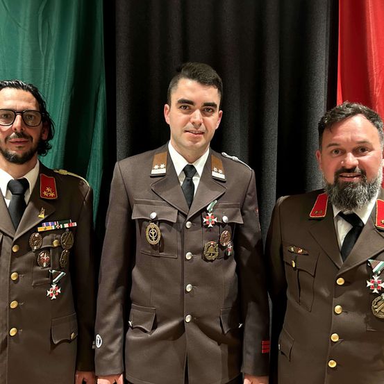 Drei Männer in Militäruniformen stehen zusammen vor einem Hintergrund aus grünen und roten Vorhängen. Der Mann in der Mitte trägt eine Medaille mit einem Stern.