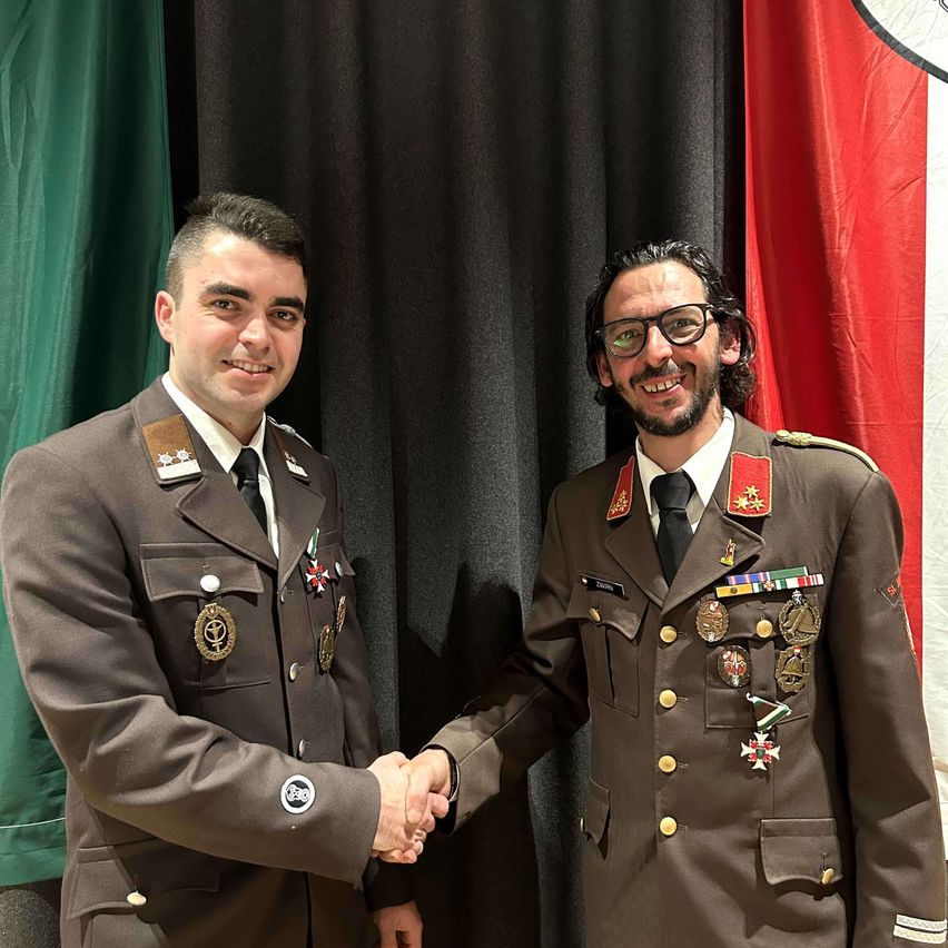 Zwei Männer in Militäruniformen schütteln sich die Hände vor einem Hintergrund mit grünen und roten Flaggen.