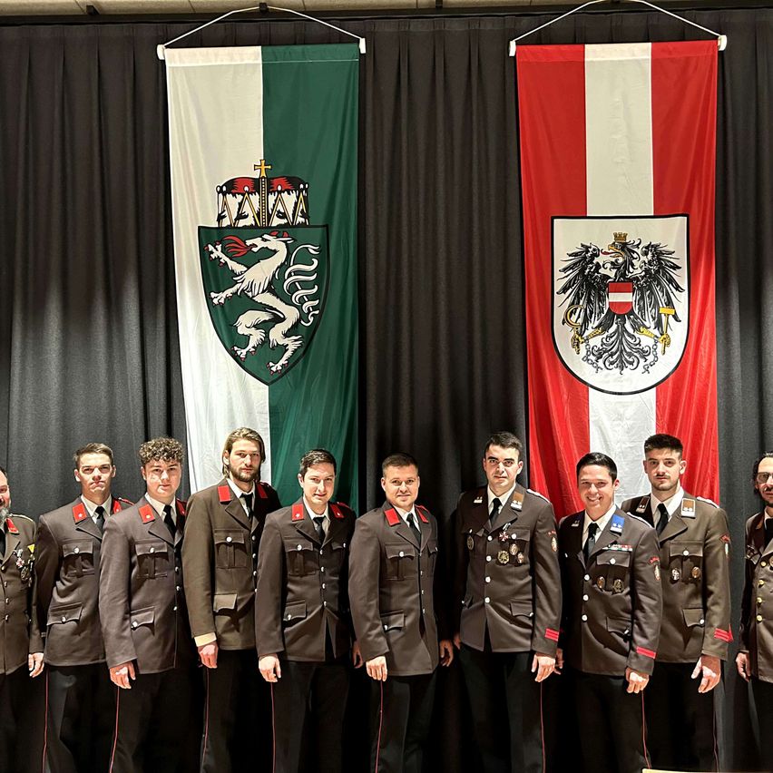 Eine Gruppe uniformierter Personen steht zusammen vor zwei Flaggen, eine mit grünem Hintergrund und weißem Schild, die andere mit rotem Hintergrund und weißem Schild.