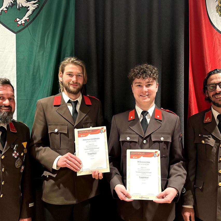 Vier Männer in Militäruniformen stehen zusammen und halten Urkunden. Der Mann in der Mitte lächelt und der Mann auf der rechten Seite trägt eine Brille. Hinter ihnen befinden sich zwei Flaggen, eine grün-weiße und eine rot-weiße.