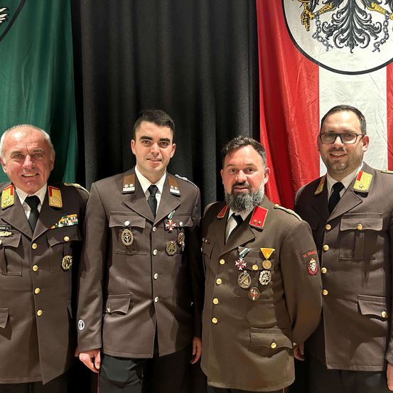 Vier Männer in Militäruniformen stehen vor einer Flagge mit einem Wappen. Ein Mann hat einen Bart und alle haben Medaillen auf der Brust.