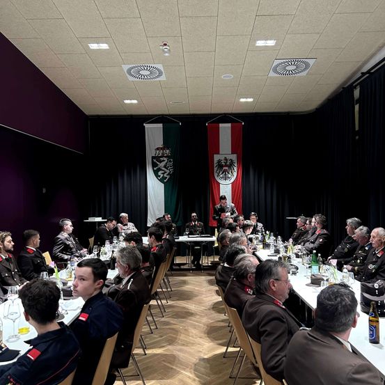 Ein Raum mit Holzboden und einem Tisch mit Flaschen und Gläsern. Es sitzen und stehen Menschen in Uniformen. Zwei Fahnen hängen an der Wand.