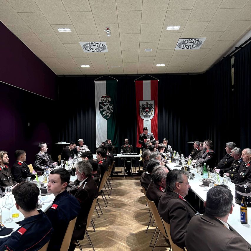 Ein Raum mit Holzboden und einem Tisch mit Flaschen und Gläsern. Es sitzen und stehen Menschen in Uniformen. Zwei Fahnen hängen an der Wand.