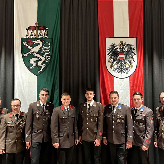 Eine Gruppe von Militäroffizieren in Uniform steht vor zwei Bannern. Ein Banner zeigt einen weißen Löwen auf rotem Hintergrund, das andere einen zweiköpfigen Adler auf rotem und weißem Hintergrund.