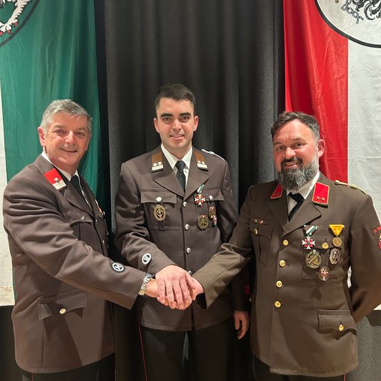 Drei Männer in Militäruniformen stehen lächelnd zusammen. Der Mann in der Mitte trägt viele Medaillen. Hinter ihnen stehen zwei Flaggen, eine grün und eine rot.