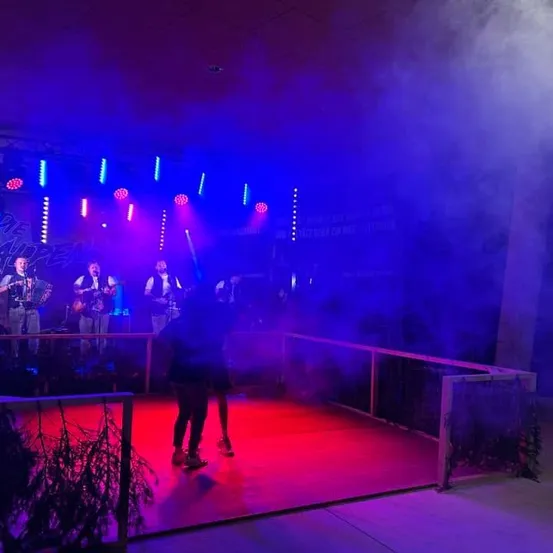 Eine Band spielt auf einer Bühne mit roter und blauer Beleuchtung. Die Bühne ist von einem Geländer umgeben, und ein Paar tanzt im Vordergrund.